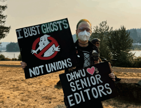 A masked protester holds two signs reading: I bust ghosts, not unions! DHWU loves Senior Editors!